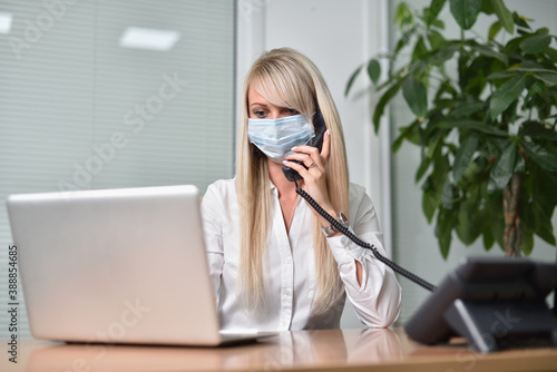 Young blonde business woman in a face mask talking on the phone in the office and working with a laptop © pridannikov