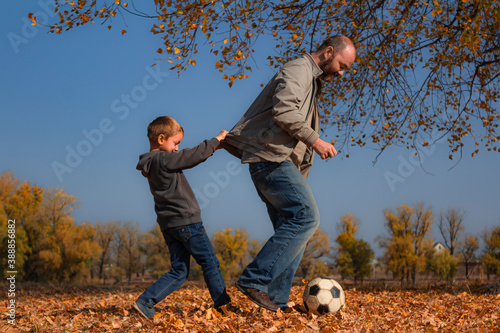 Father and son play a soccer ball outdoors on a sunny day among the autumn foliage.