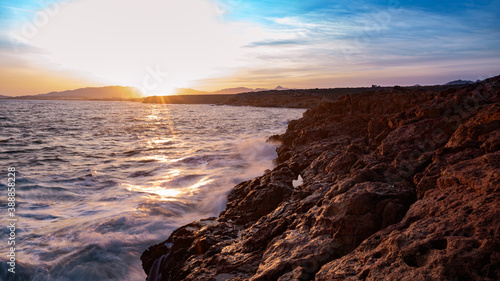 Ocean landscape, rocks next to the sea and the waves crashing in the red rocks. Sunshine in the sunset with blue sky and the sun reflections on the sea.