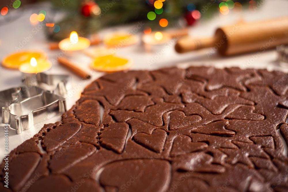 Brown chocolate dough, shaped in cookies of various form, lays on the table. Christmas decoration on the background. - obrazy, fototapety, plakaty 