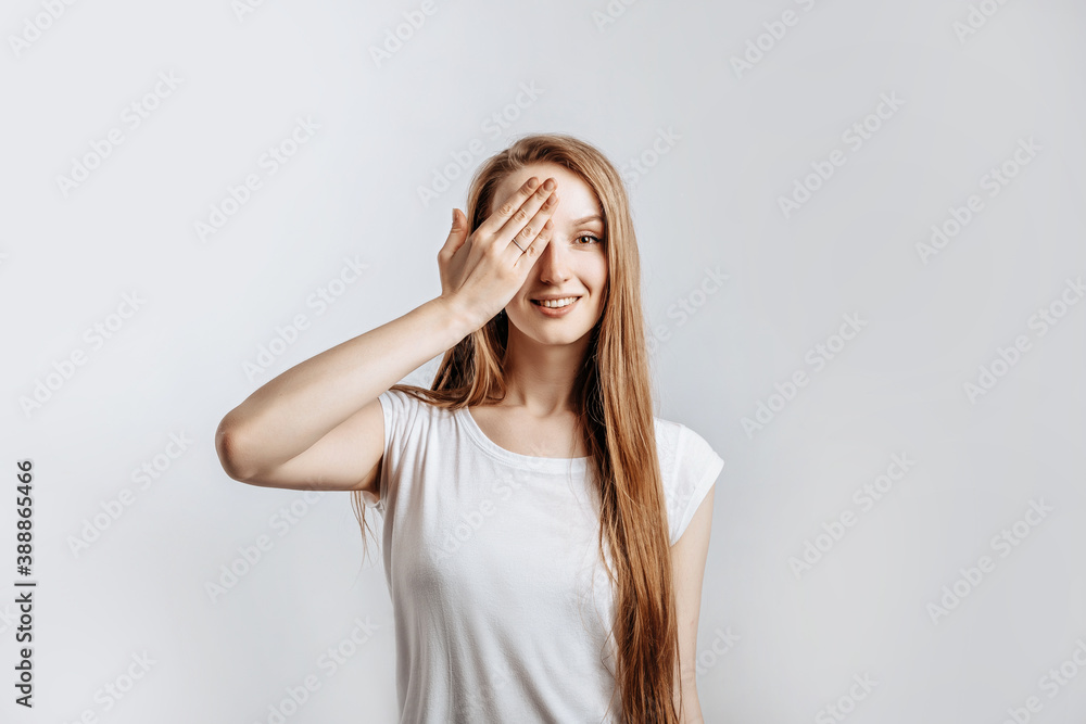 Young beautiful girl smiling and covering half of her face with hand on gray isolated background