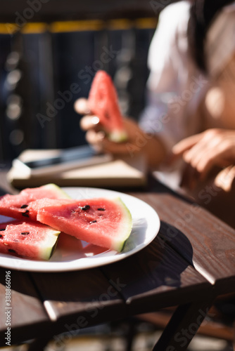 blurred view of woman eating watermelon on balcony and plate with watermelon slices on foreground