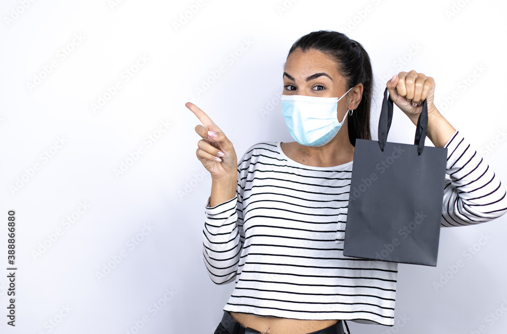 Young beautiful woman holding a black shopping bag wearing a medical mask to protect herself from the coronavirus pointing to the side . Black Friday Young beautiful woman holding a black shopping bag wearing a medical mask to protect herself from the coronavirus pointing to the side . Black Friday