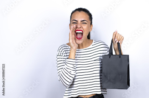 Young beautiful woman holding a black shopping bag shouting and screaming loud to side with hands on mouth. Black Friday