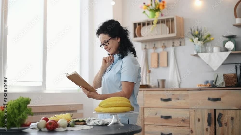 Woman with black curly hair standing in the kitchen and reading book.