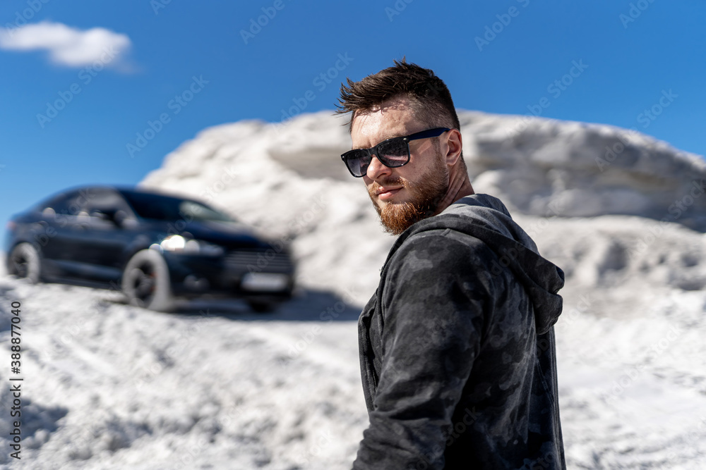 Portrait of a stylish bearded man in trendy sunglasses and leather jacket stands near black car. Fashion portrait on white rocks background. Portrait of a stylish bearded man in trendy sunglasses and leather jacket stands near black car. Fashion portrait on white rocks background.