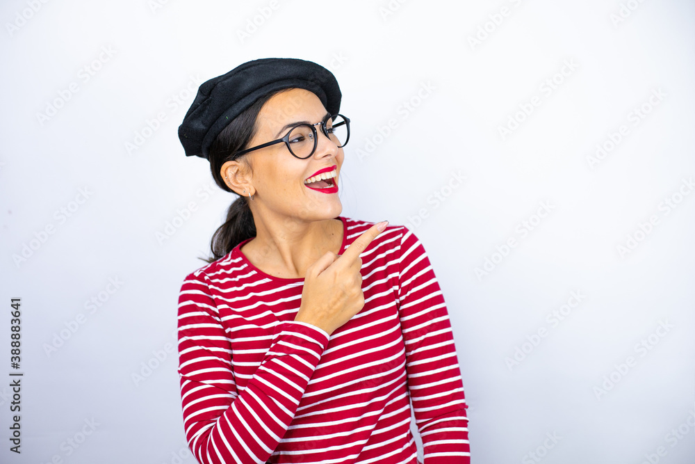 Young beautiful brunette woman wearing french beret and glasses over white background surprised and pointing with hand and finger to the side Young beautiful brunette woman wearing french beret and glasses over white background surprised and pointing with hand and finger to the side