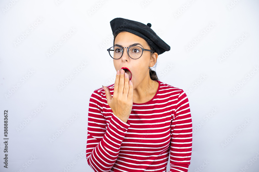 Young beautiful brunette woman wearing french beret and glasses over white background with her hand over her mouth and surprised, looking side