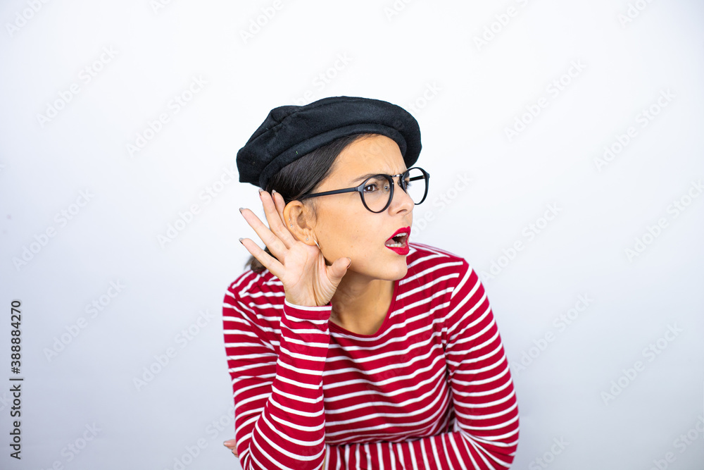 Young beautiful brunette woman wearing french beret and glasses over white background surprised with hand over ear listening an hearing to rumor or gossip Young beautiful brunette woman wearing french beret and glasses over white background surprised with hand over ear listening an hearing to rumor or gossip