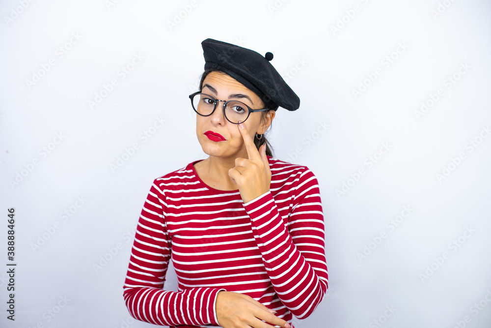 Young beautiful brunette woman wearing french beret and glasses over white background Pointing to the eye watching you gesture, suspicious expression Young beautiful brunette woman wearing french beret and glasses over white background Pointing to the eye watching you gesture, suspicious expression
