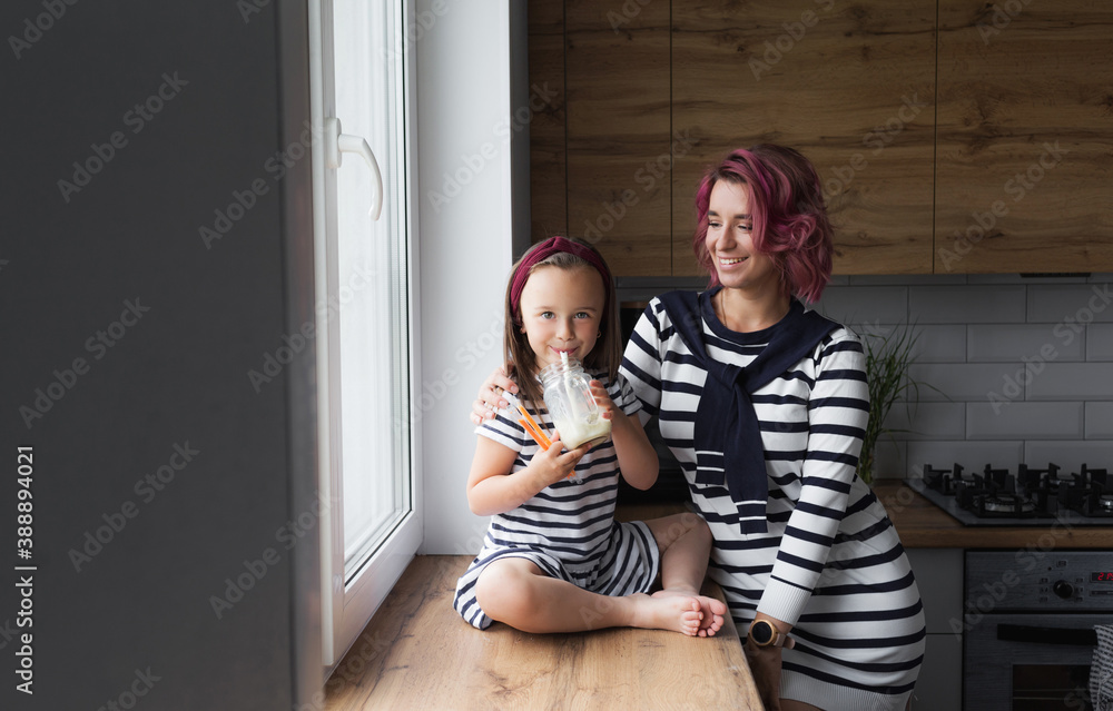 happy family, mother and her little daughter are sitting on the windowsill in the kitchen and drinking milk. They are dressed in beautiful dresses with black and white stripes
