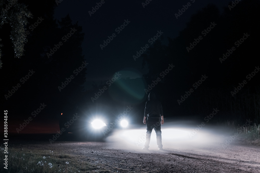A mysterious hooded figure standing in front of a car at night A mysterious hooded figure standing in front of a car at night