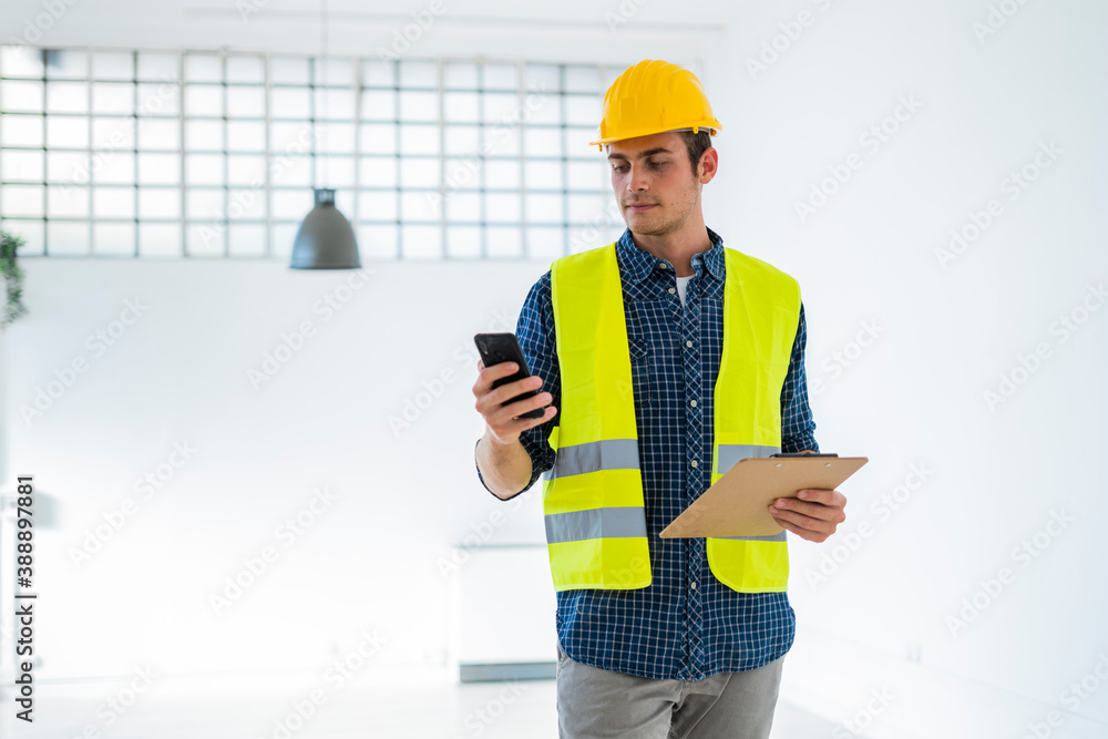 Young man wearing in work helmet and jacket using mobile phone while standing at construction site Young man wearing in work helmet and jacket using mobile phone while standing at construction site