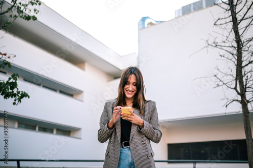 Smiling woman text messaging on smart phone while standing against building exterior photo