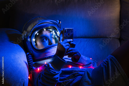 Boy wearing space helmet using mobile phone while relaxing on sofa in illuminated home