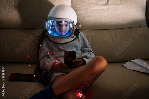 Boy wearing space helmet using smart phone while sitting with illuminated lighting equipment on sofa photo