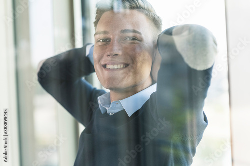 Close-up of smiling businessman with hands behind head seen through window in office photo