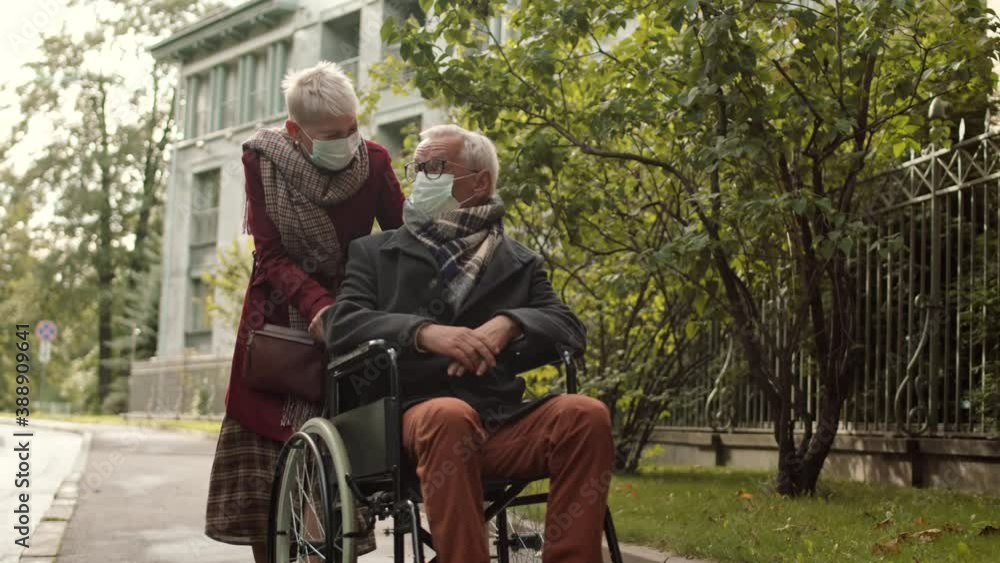 Full shot of senior cheerful Caucasian woman walking with her handicapped husband on wheelchair. Couple wearing masks, walking down street and laughing Full shot of senior cheerful Caucasian woman walking with her handicapped husband on wheelchair. Couple wearing masks, walking down street and laughing