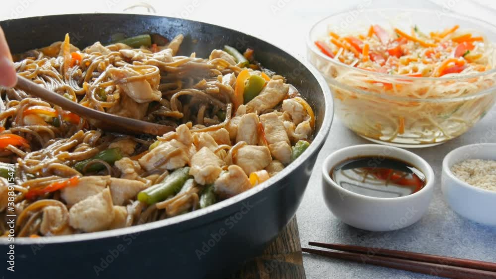 Japanese soba rice noodles with pieces of chicken, carrot, green shoots, sesame seeds, soy sauce. National food in a restaurant in large frying pan next to wooden or bamboo fork. Glass noodle salad. Japanese soba rice noodles with pieces of chicken, carrot, green shoots, sesame seeds, soy sauce. National food in a restaurant in large frying pan next to wooden or bamboo fork. Glass noodle salad.