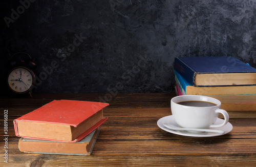 White coffee cup with dark black coffee And old books And the clock is arranged on an old wooden table and a black wooden wall.