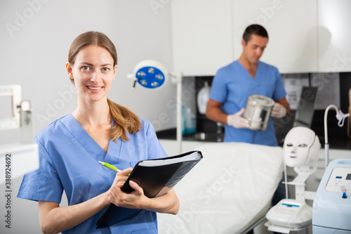 Positive female doctor cosmetologist wearing blue overall meeting client while male nurse preparing equipment before procedure