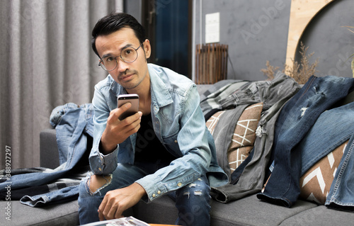 Man wearing denim jacket and jean with holding smartphone and looking during sitting on sofa in modern living room at home , Jean fashion men concept