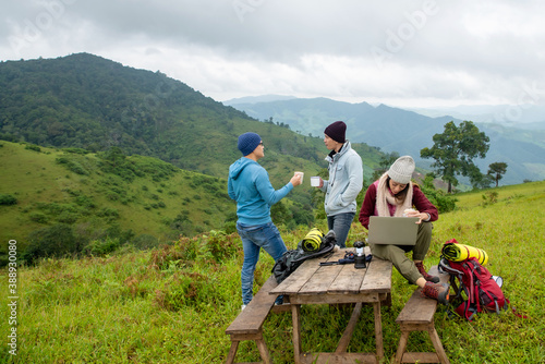 Group of Happy Asian man and woman friends backpacker enjoy hiking and drinking coffee together on forest mountain in autumn. Young woman using laptop computer online working in outdoor vacation
