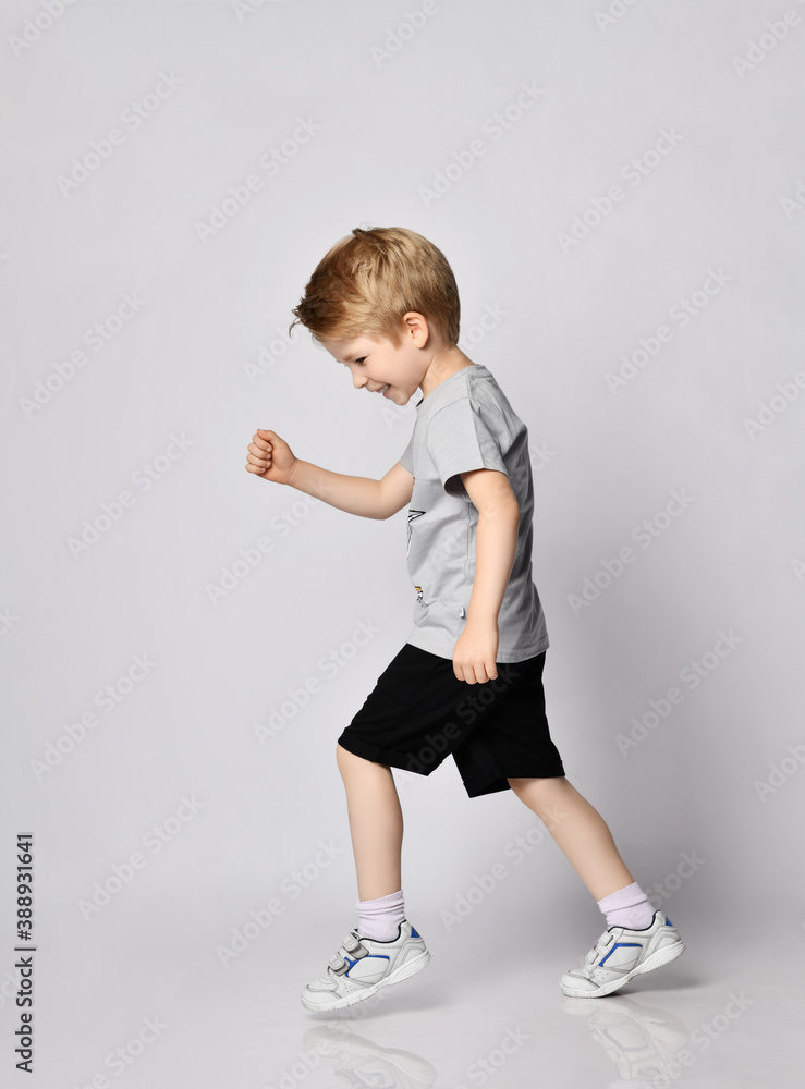 Playful smiling active blond kid boy in t-shirt and black shorts walks side to camera towards copy space and looks down Playful smiling active blond kid boy in t-shirt and black shorts walks side to camera towards copy space and looks down
