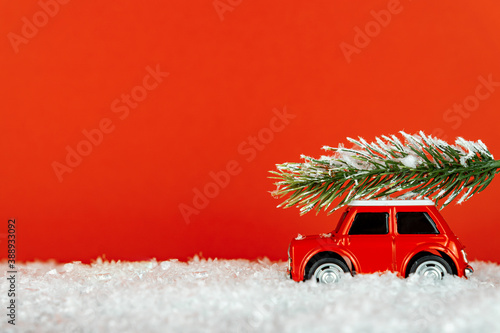 Small red toy car carrying spruse tree on a snowy road. Festive christmas greeting card.