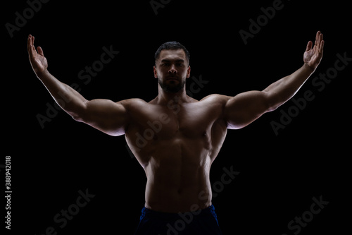 Portrait of a bodybuilder standing isolated on black background in a shadow with raised hands to show off his muscles