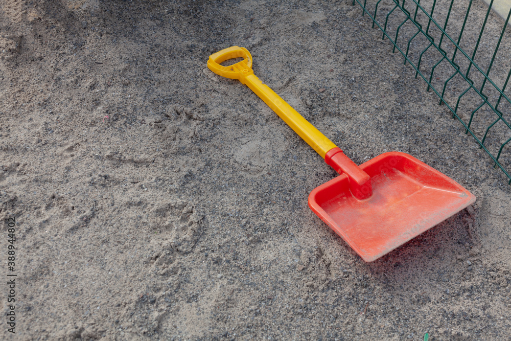 Yellow and red shovel on the playground lies on the sand near the green fence. Yellow and red shovel on the playground lies on the sand near the green fence.