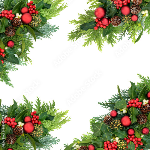 Christmas abstract border for the festive season with red bauble decorations & winter greenery of holly, ivy, mistletoe, cedar cypress & pine cones on white background. Top view, flat lay, copy space.