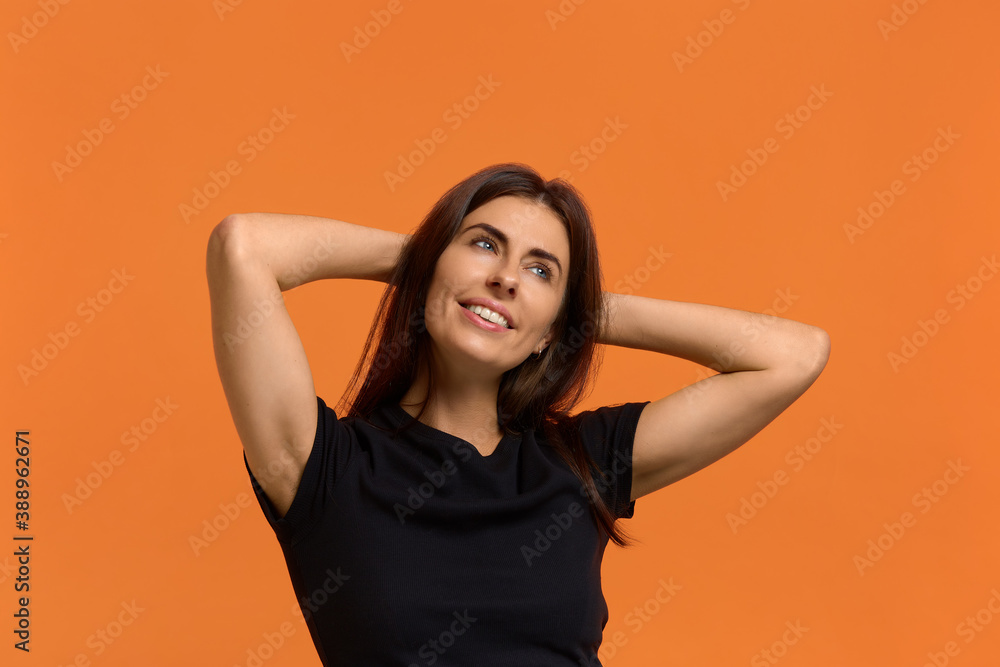 What a wounderful life. Relaxed carefree caucasian woman in black t-shirt keeps hands behind head, has positive expression, smiles pleasantly, enjoys holidays. Isolated over an orange background. What a wounderful life. Relaxed carefree caucasian woman in black t-shirt keeps hands behind head, has positive expression, smiles pleasantly, enjoys holidays. Isolated over an orange background.