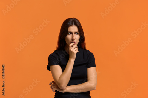 Hmm. I need to think. Suspicious thoughtful caucasian woman in black t-shirt keeping hand on her face as if trying hard to figure out what to do. Isolated over an orange background.