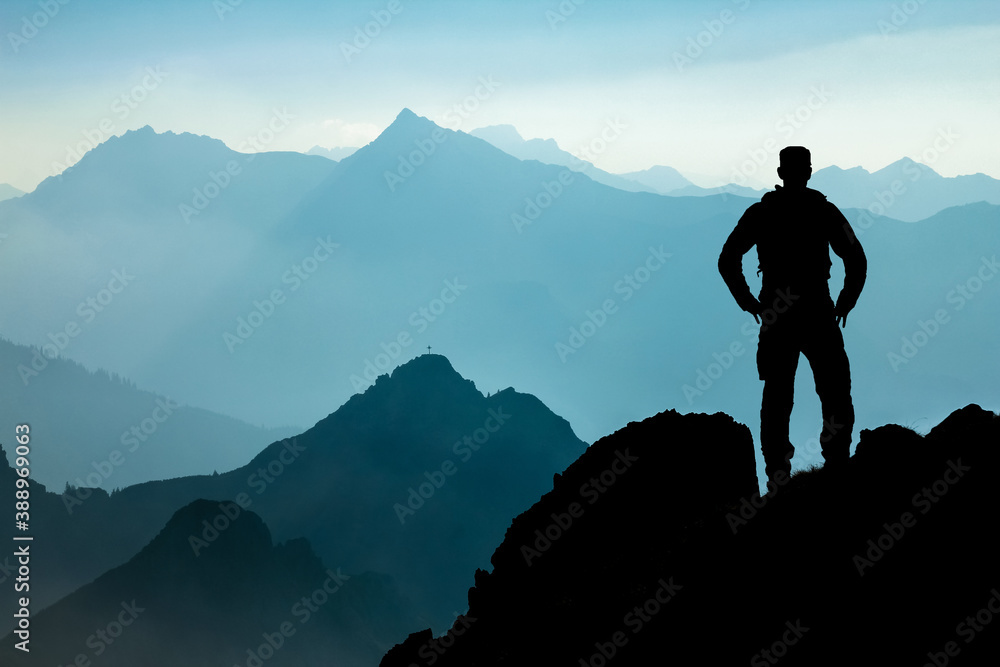 One Man reaching summit after climbing and hiking enjoying freedom and looking towards mountains silhouettes panorama during sunrise.