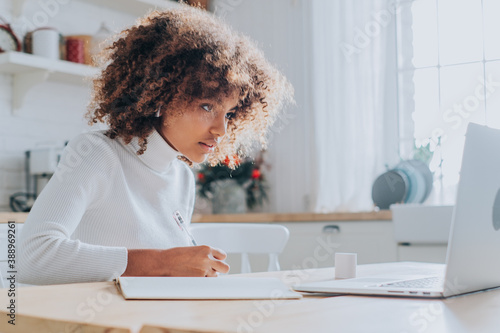Fotografie An African-American student with a buxom hairstyle watches and notes a lecture online on her laptop at home in the kitchen on a Sunny day
