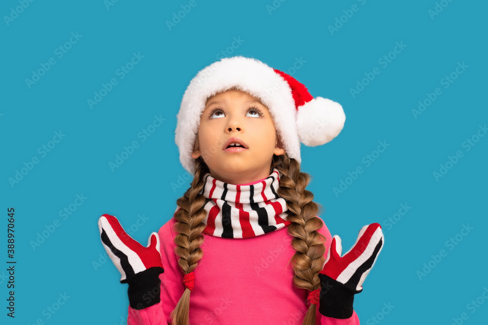 A little girl in a Christmas hat looks up in surprise on a blue background. A little girl in a Christmas hat looks up in surprise on a blue background.