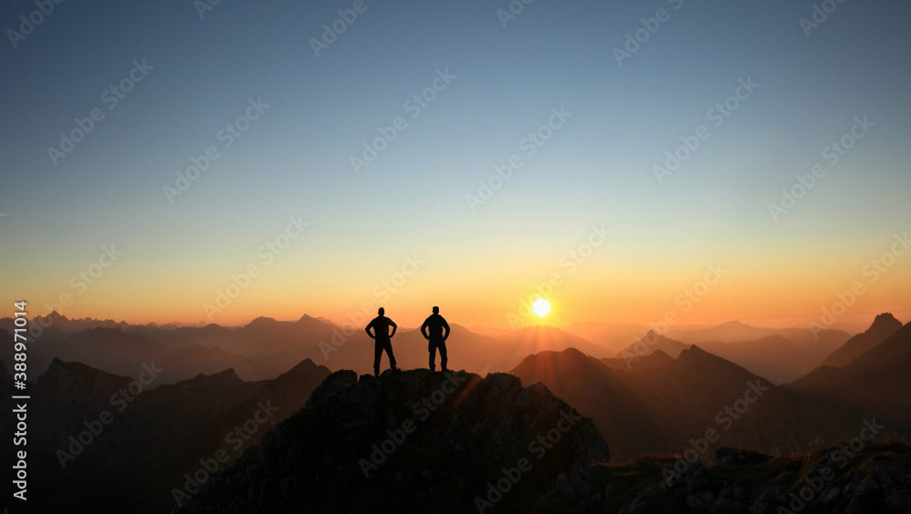 Two Men reaching summit enjoying freedom and looking towards mountains sunset. - obrazy, fototapety, plakaty 