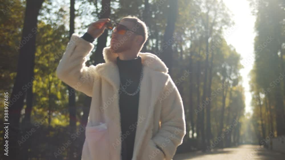 Portrait of fashionable young man wearing white faux fur coat and sunglasses walking in fall park Portrait of fashionable young man wearing white faux fur coat and sunglasses walking in fall park