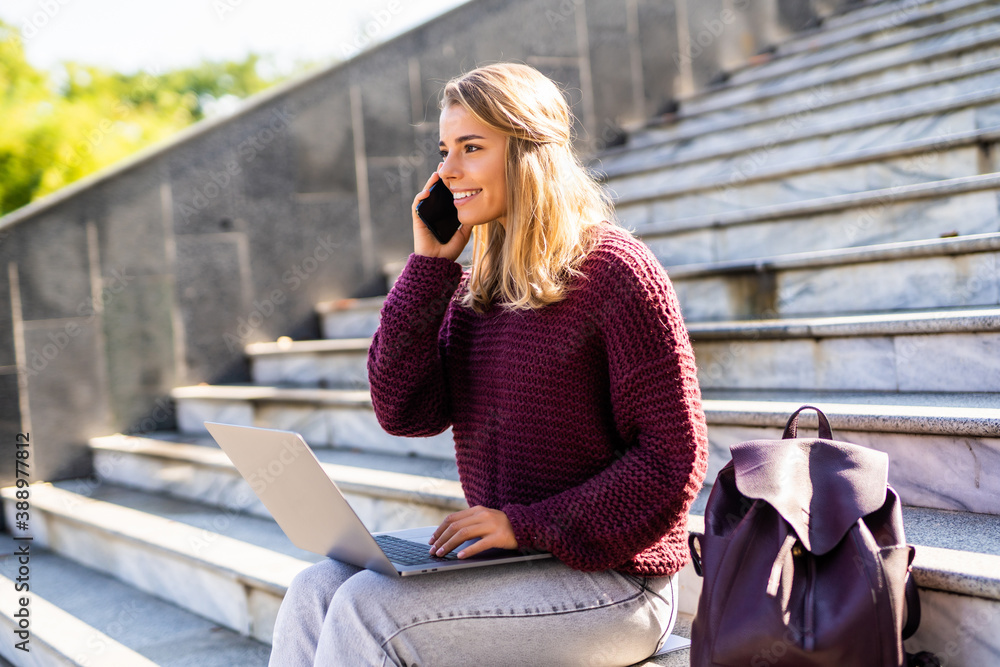 Fototapeta premium Happy young woman talking on mobile phone while sitting outdoors with laptop