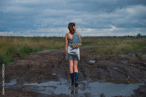 Fashion model woman holding string bag with potatoes, stay in dirty puddle in field, she is wearing a silver short dress and blue rubber boots. Concept of village style.