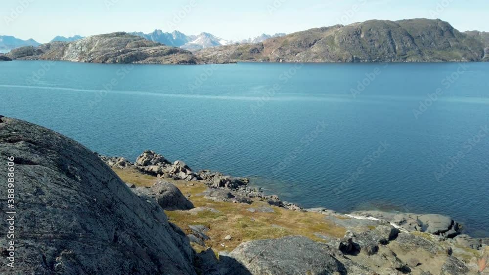 Scenic view of rocky shoreline along coast of Greenland from top of rocks lining the beach. TILT UP reveals distant snow capped mountains on opposite side of fjord, calm blue water and clear sky. Scenic view of rocky shoreline along coast of Greenland from top of rocks lining the beach. TILT UP reveals distant snow capped mountains on opposite side of fjord, calm blue water and clear sky.
