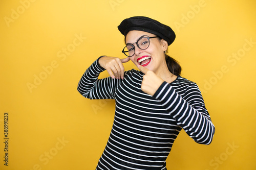 Young beautiful brunette woman wearing french beret and glasses over yellow background doing the “call me” gesture with her hands.