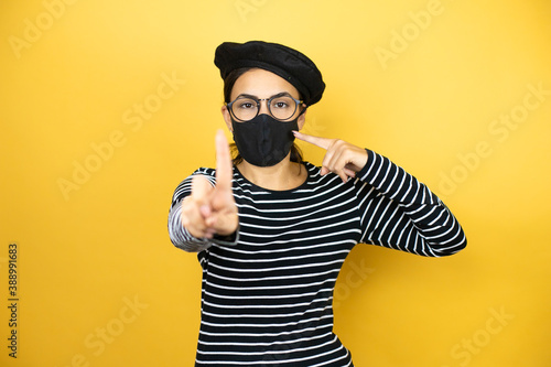 Young beautiful brunette woman wearing french beret and glasses over yellow background pointing the mask. Warning expression with negative and serious gesture on the face.