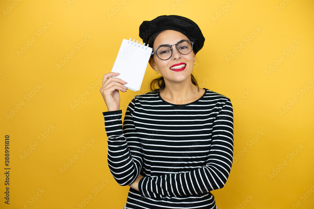 Young beautiful brunette woman wearing french beret and glasses over yellow background smiling and showing blank notebook in her hand Young beautiful brunette woman wearing french beret and glasses over yellow background smiling and showing blank notebook in her hand
