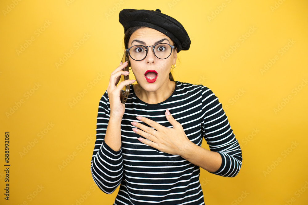 Young beautiful brunette woman wearing french beret and glasses over yellow background talking on the phone with a worried expression Young beautiful brunette woman wearing french beret and glasses over yellow background talking on the phone with a worried expression