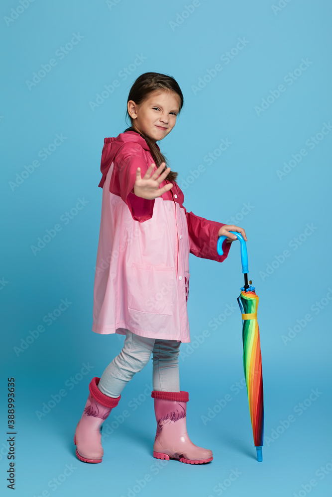 cute child girl with multicolored umbrella in pink rain coat and rubber boots showing stop gesture on blue background. cute child girl with multicolored umbrella in pink rain coat and rubber boots showing stop gesture on blue background.