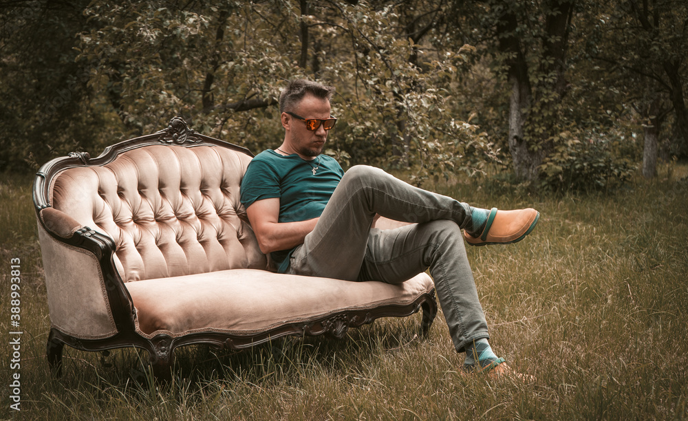 Obraz premium Resting man in grey jeans sitting on a sofa in the middle of garden. High quality photo