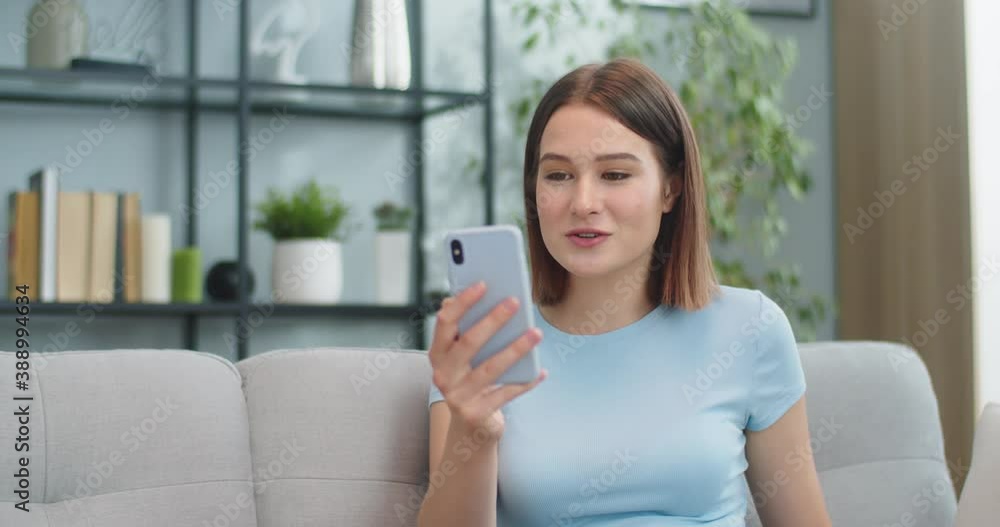 Charming Caucasian woman sitting on sofa and talking to family via video conference. Attractive young female using smarphone to call in living room at home. Self-isolation, communication concept. Charming Caucasian woman sitting on sofa and talking to family via video conference. Attractive young female using smarphone to call in living room at home. Self-isolation, communication concept.