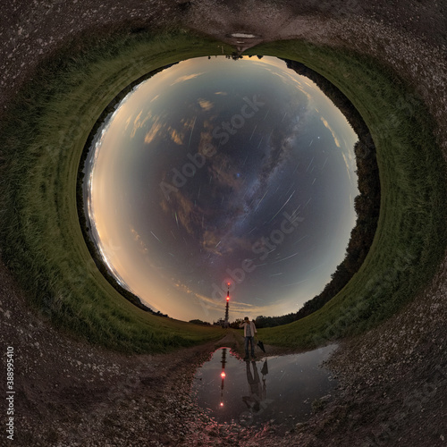 360 degrees panorama of the full sky, a man standing on the road looks at the reflection in the water under a night sky full of meteors of the Perseids and the Milky Way.